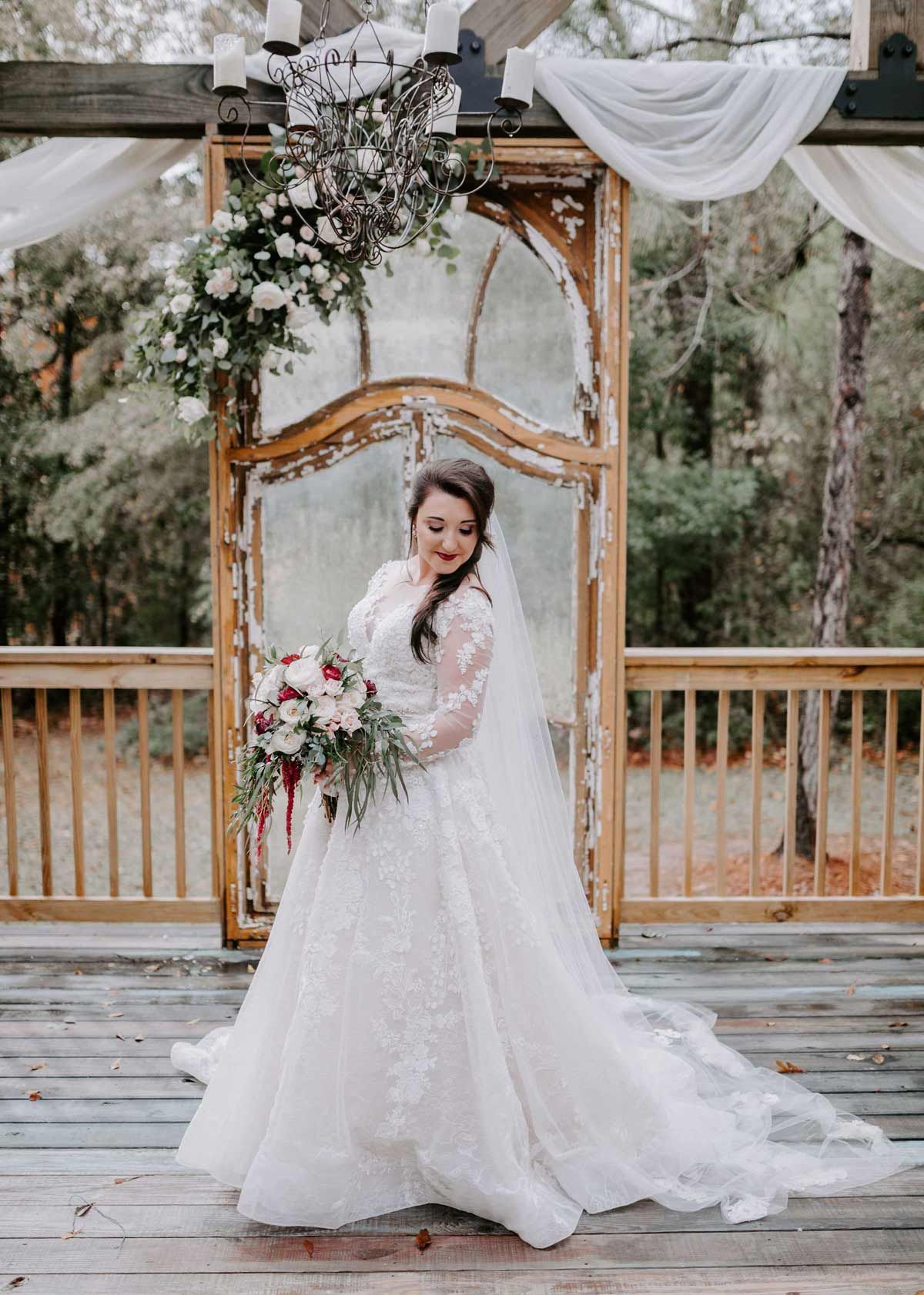 Bride in lace gown holding bouquet, standing on wooden deck in front of ornate archway.