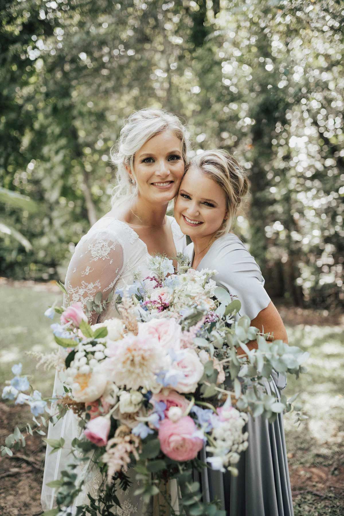 Bride and bridesmaid, smiling, holding large floral bouquet in a garden setting.