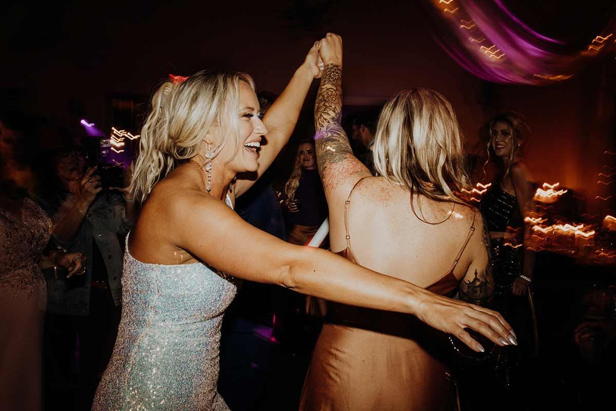 Two women dancing at a party; one with a beaded dress and the other with a backless dress, both with arms raised.