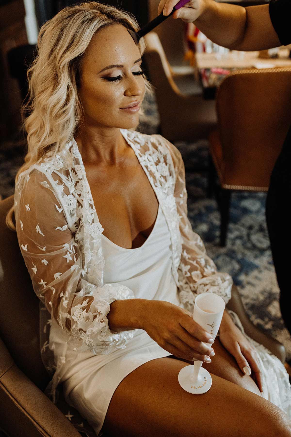 Woman in white robe, having hair styled, holding champagne flute.