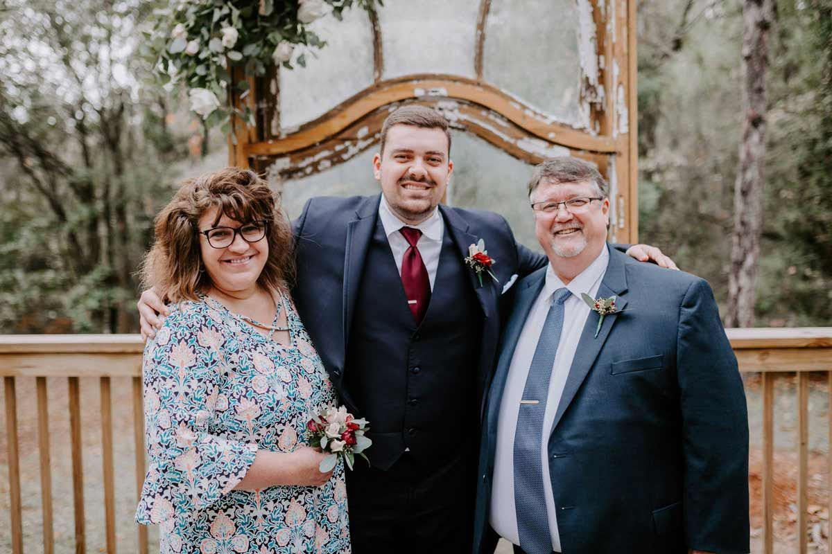 Family smiling together outdoors; man in suit flanked by parents.