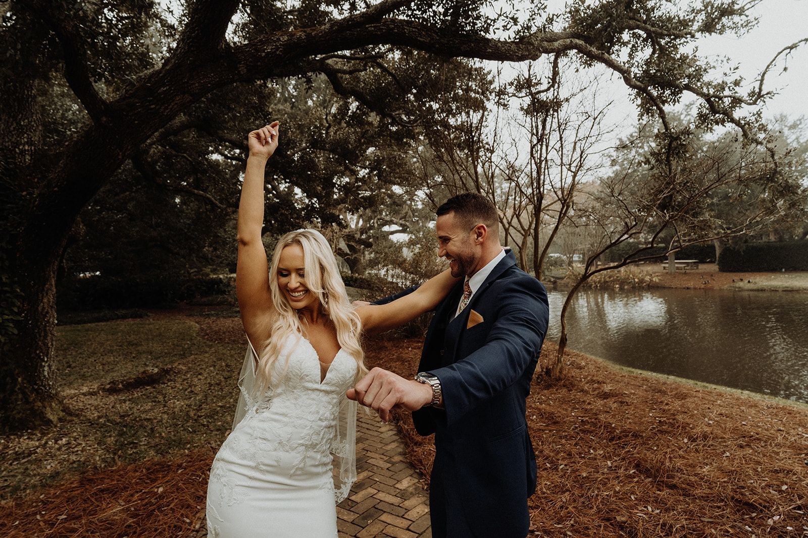 Bride celebrating with arm raised as groom holds her hand; they are outside near a pond.