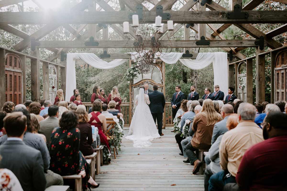 Wedding ceremony in a wooden gazebo; couple at the altar, guests seated, draped fabric and greenery overhead.