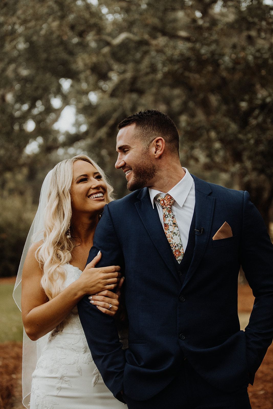 Bride and groom smile at each other; she holds his arm. They are outdoors, with a blurred natural background.