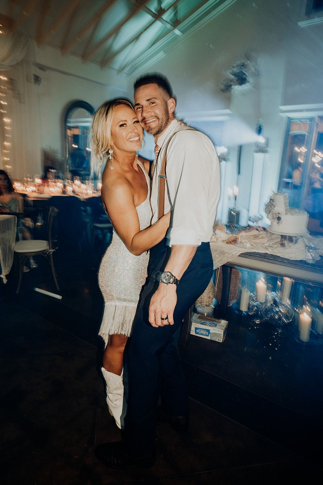 Couple dancing at a wedding reception. Woman in a white dress and boots hugs the man in a white shirt and suspenders.