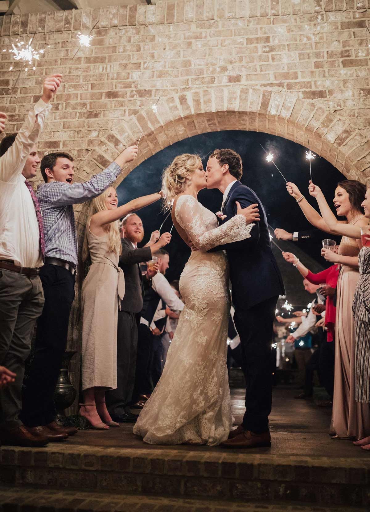 Newlyweds kiss under a brick arch as guests wave sparklers at their wedding.