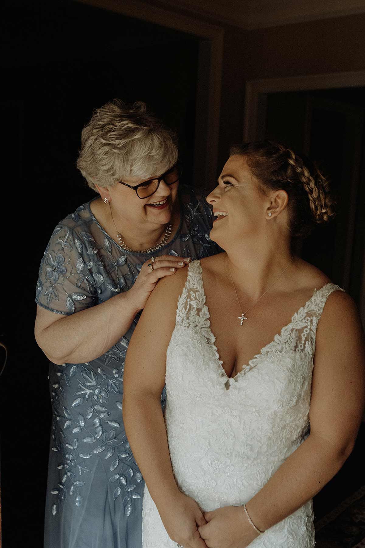 Woman in wedding dress smiles at woman in beaded dress, in a room.