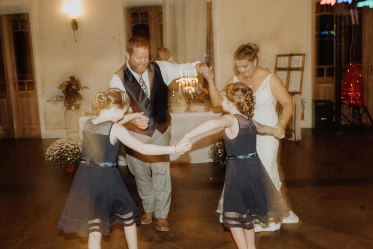 Wedding dance with a bride, groom, and two young girls in navy dresses, all holding hands and smiling.