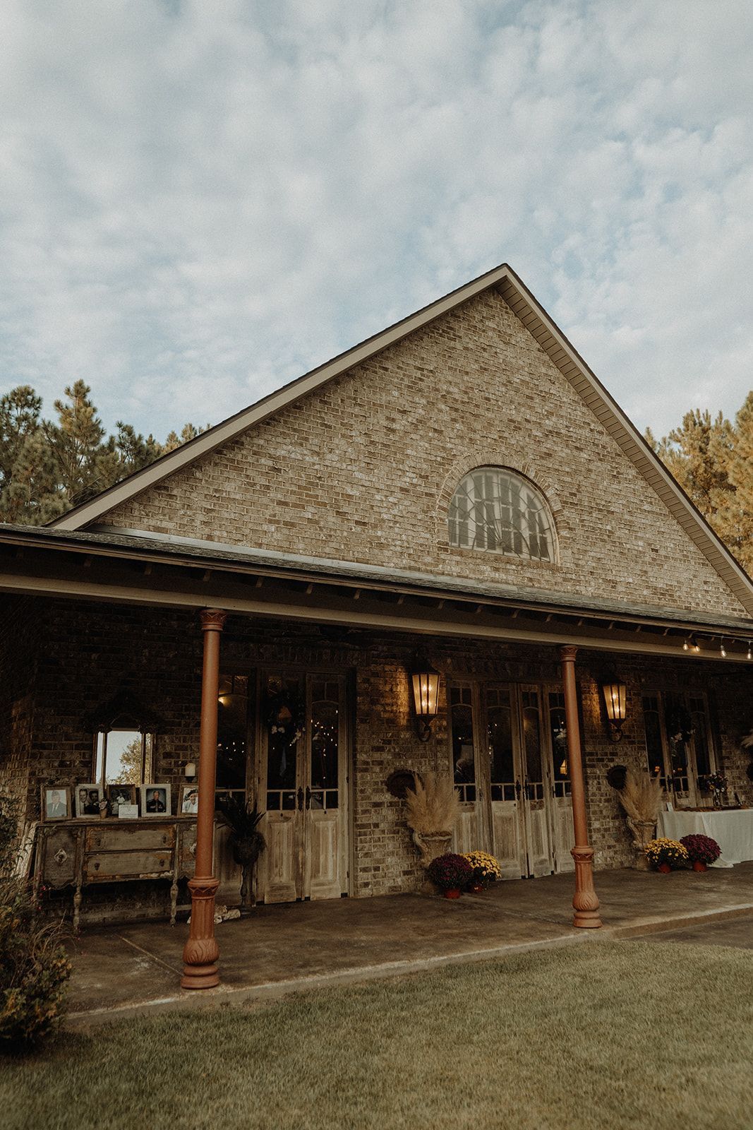 Stone building with a covered porch. Wooden doors, red columns, and arched window under a gabled roof.