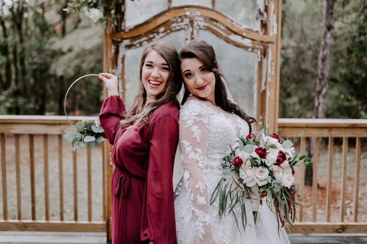 Bride in white dress and bridesmaid in maroon dress pose back-to-back, smiling, outdoors.