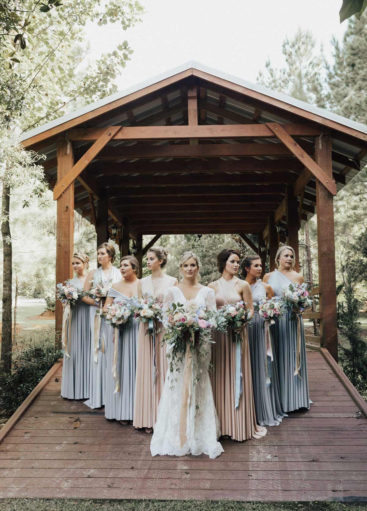 Bridesmaids in pastel gowns with the bride in front, under a wooden gazebo with flowers.