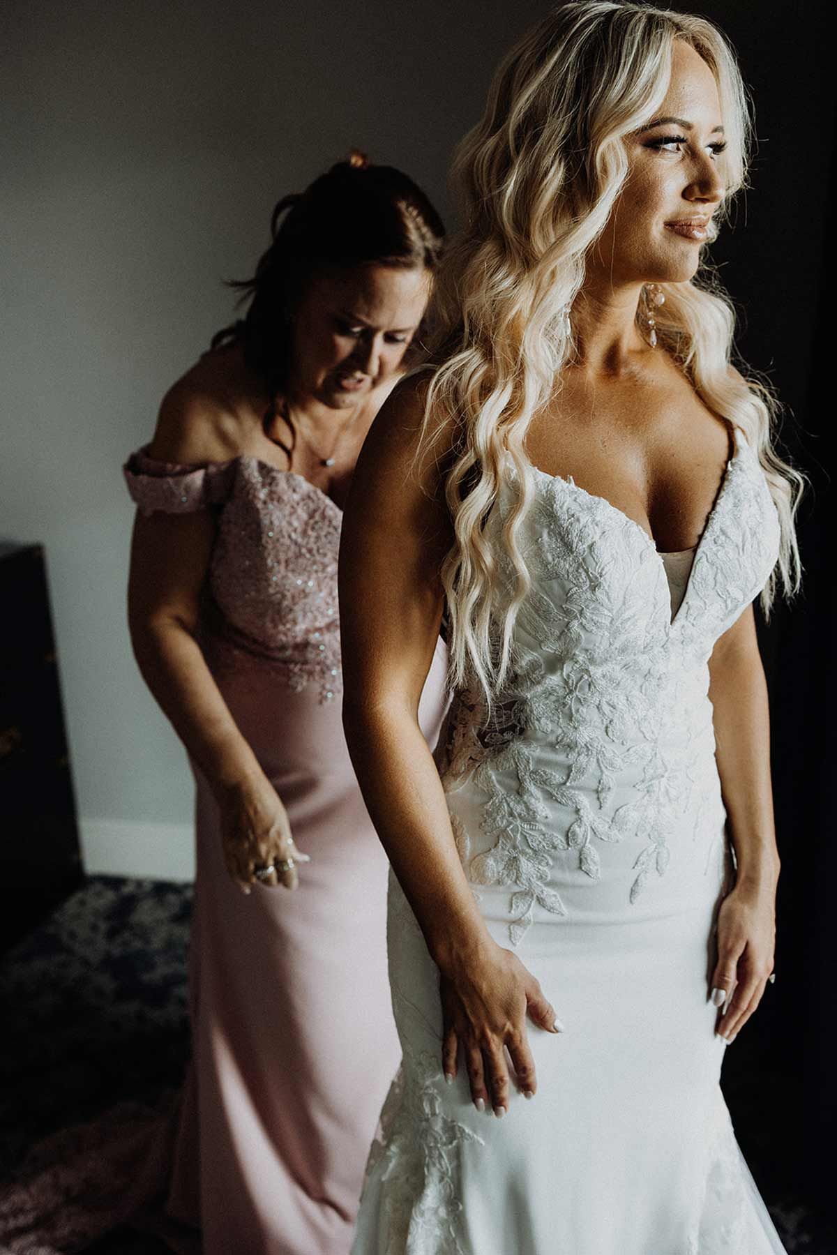 Woman in a white wedding dress, being helped by another woman in a pink dress.