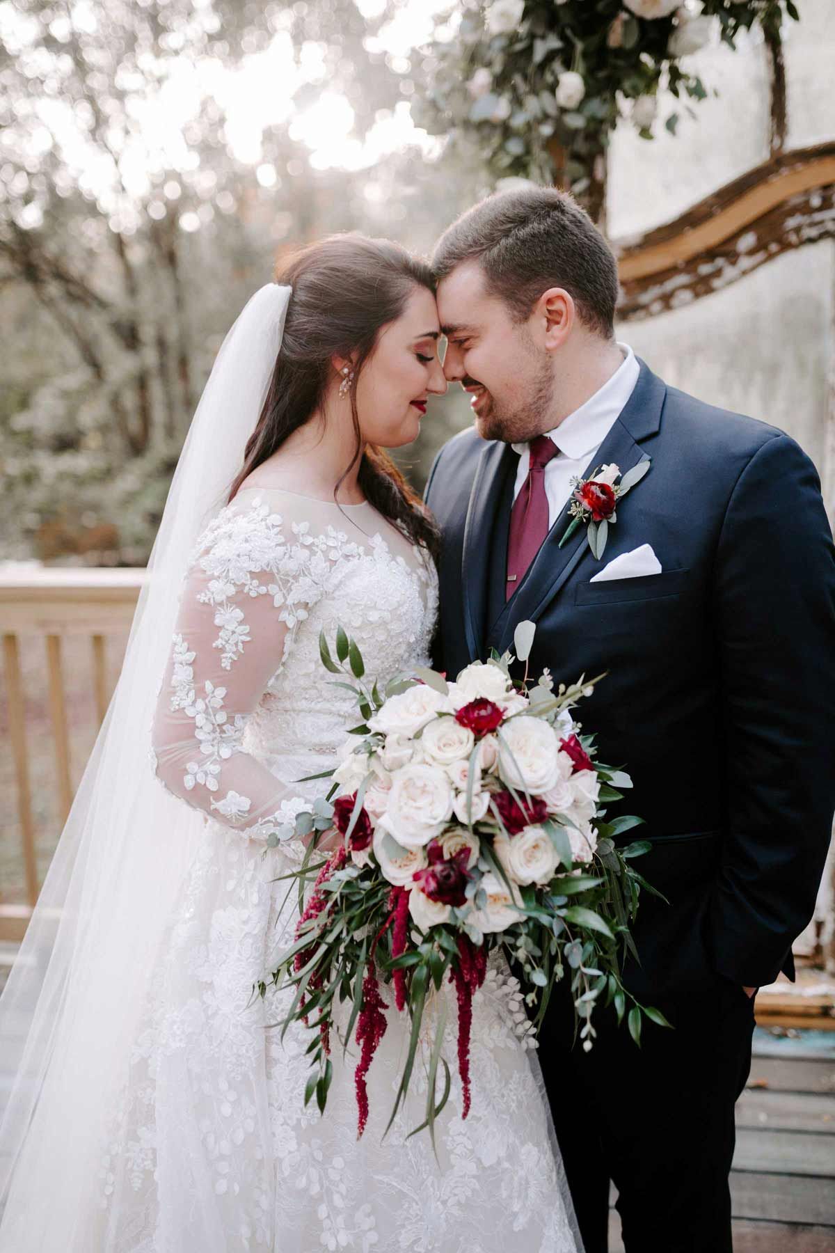 Bride and groom embrace, forehead to forehead. She wears a white gown, he a navy suit. They stand outside, holding a bouquet.