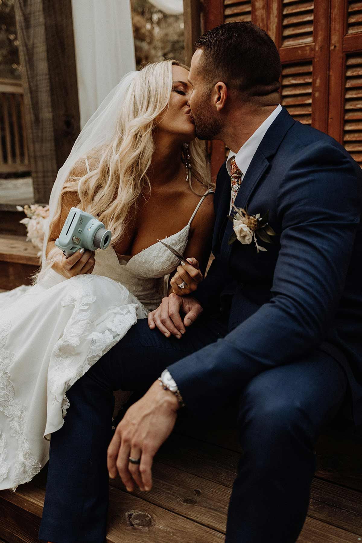 Bride and groom kissing on wooden steps, she holds a camera, he wears a navy suit.