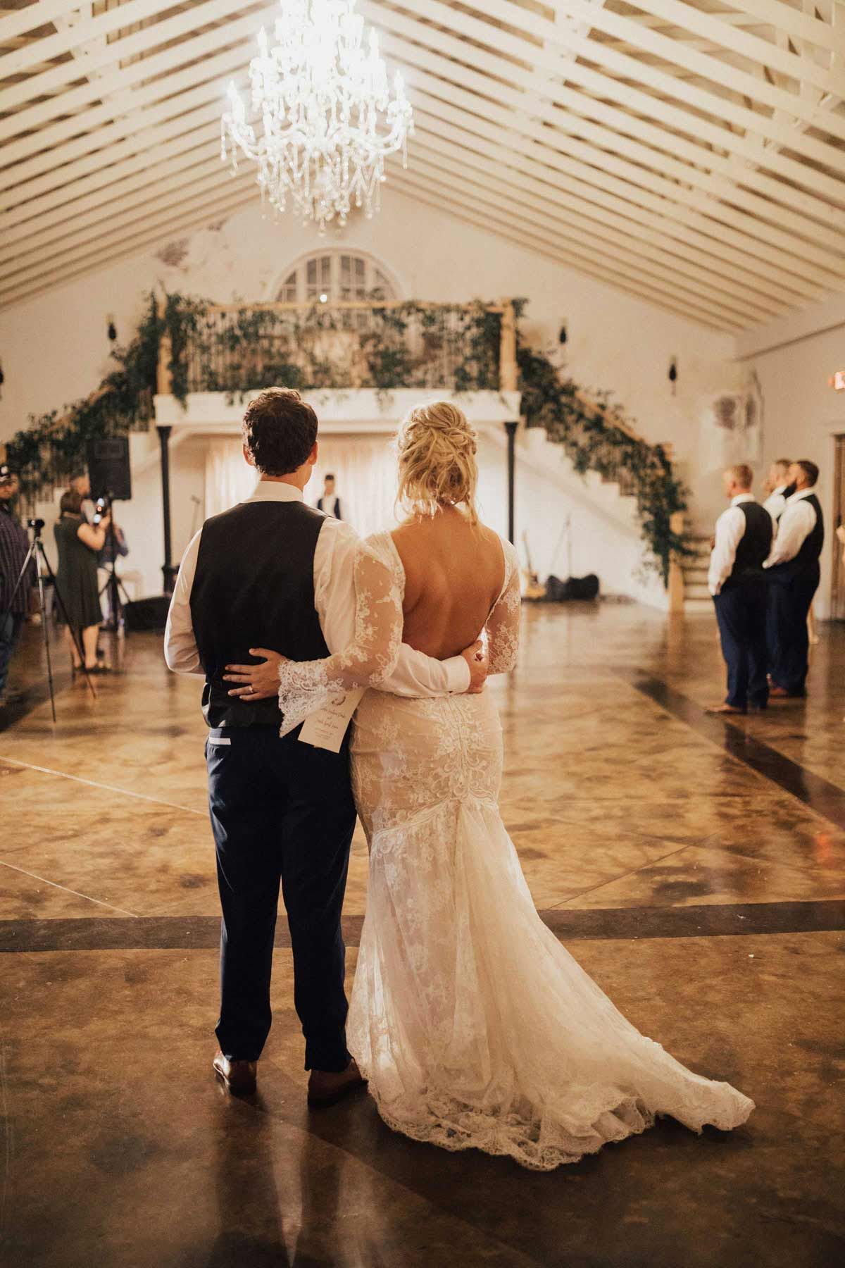 Couple in wedding attire embrace at reception; ornate chandelier above.