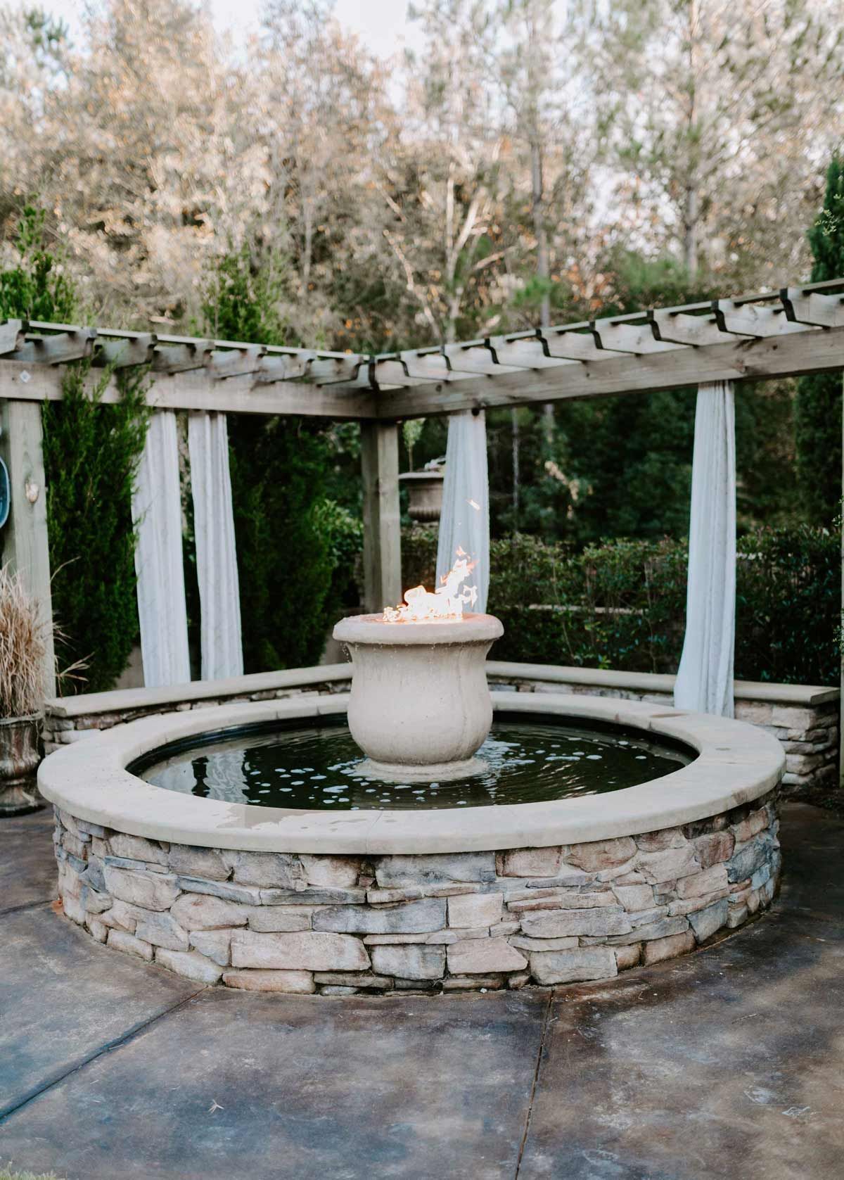 Stone fountain with pergola, white curtains, and surrounding greenery.