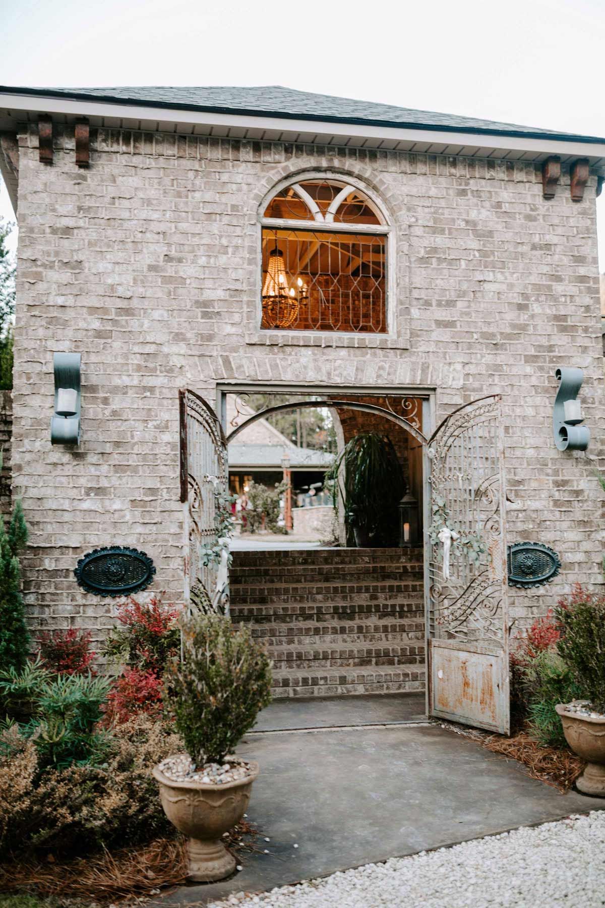 Brick building with open ornate metal gates leading to steps. Plants and light fixtures surround.