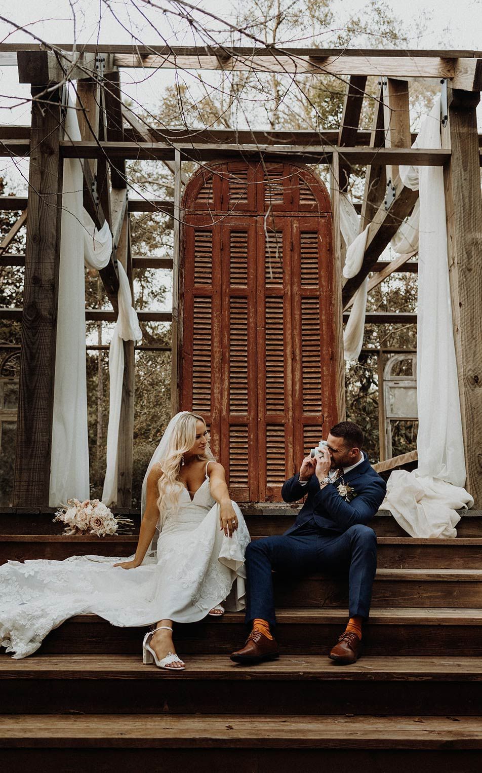 Bride and groom sit on wooden steps. Bride in white dress, groom in blue suit, both facing each other, near a weathered door.