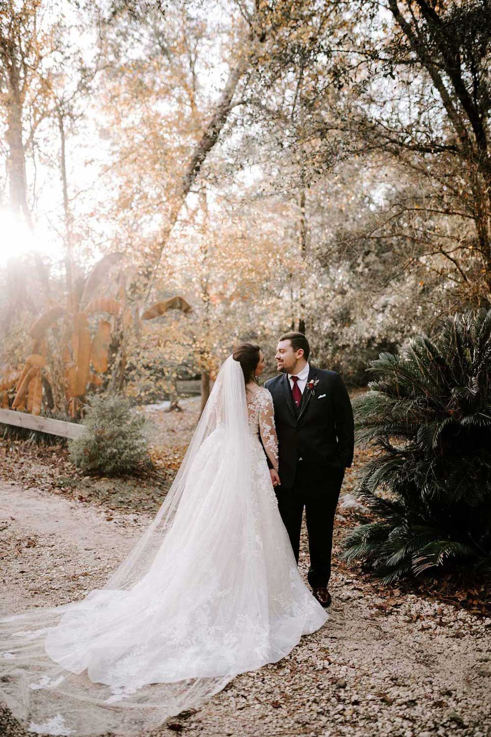 Bride and groom holding hands in a park, sunlight shining. The bride wears a white gown with a long veil.