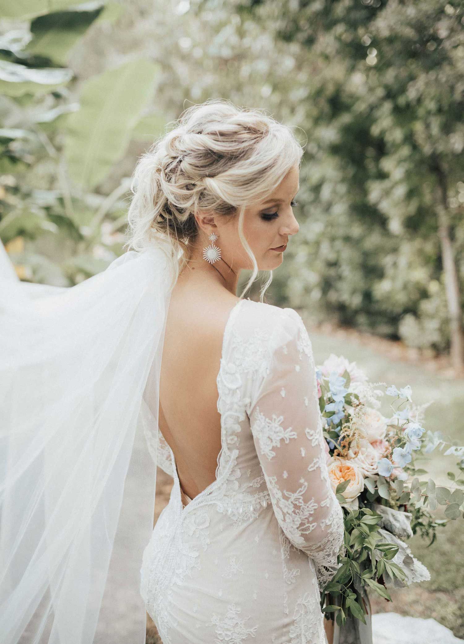 Blonde bride in white lace gown, backless design, veil, holds bouquet, looking down, outdoor setting.