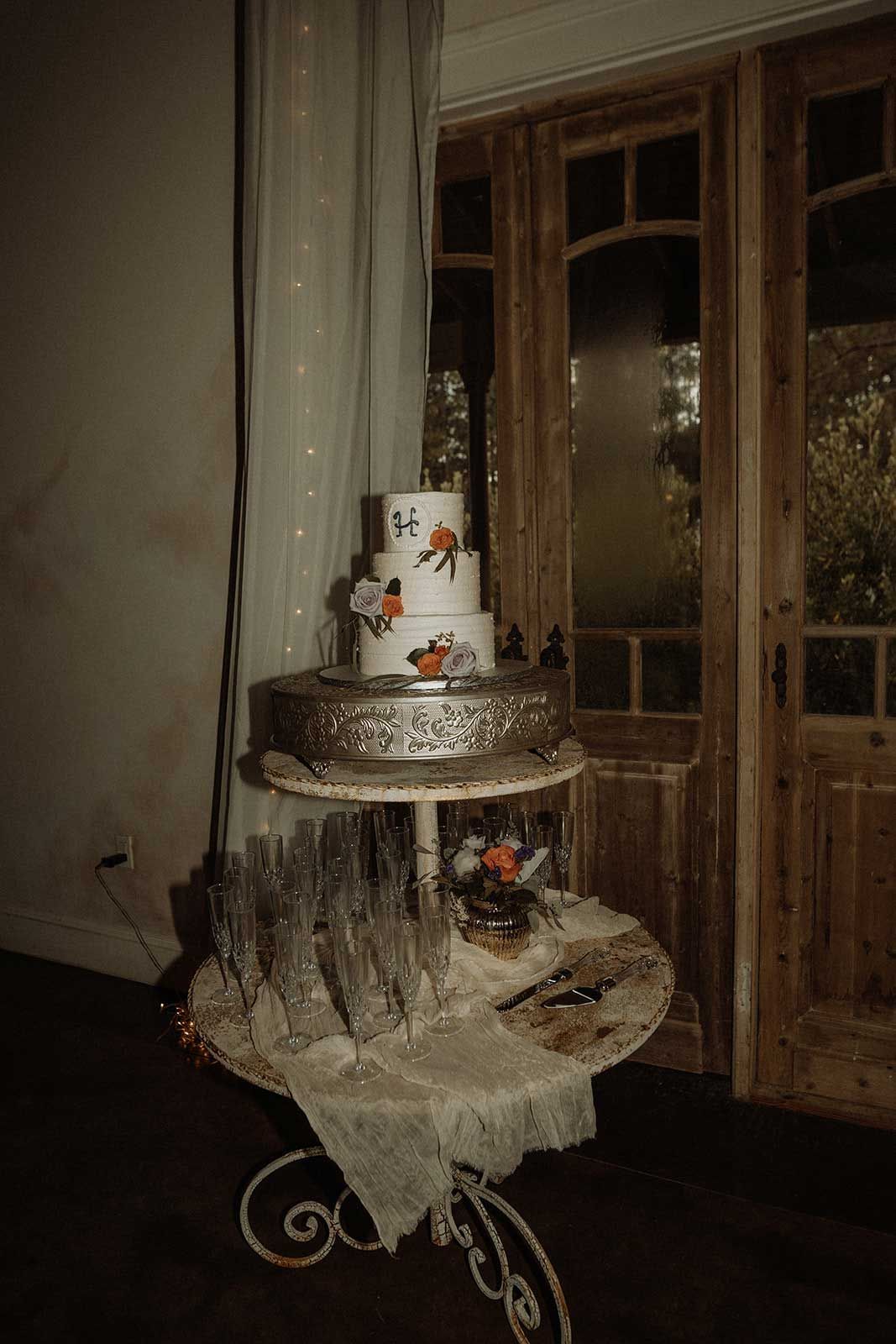 Three-tiered wedding cake on a decorative table with champagne flutes, near a window and rustic doors.