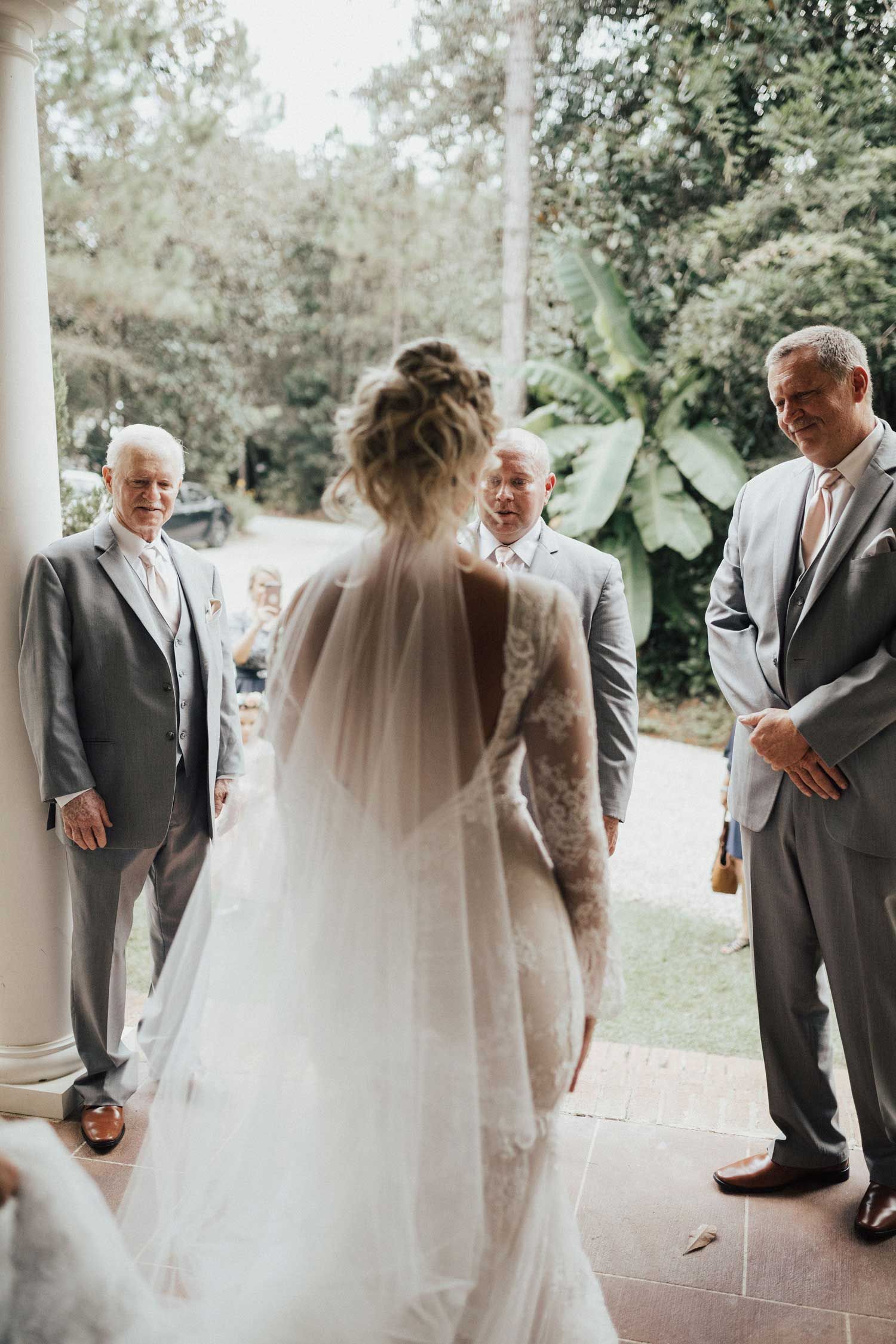 Bride in wedding dress facing three men, on a porch. Men in grey suits. Bride's back turned, veil flowing.