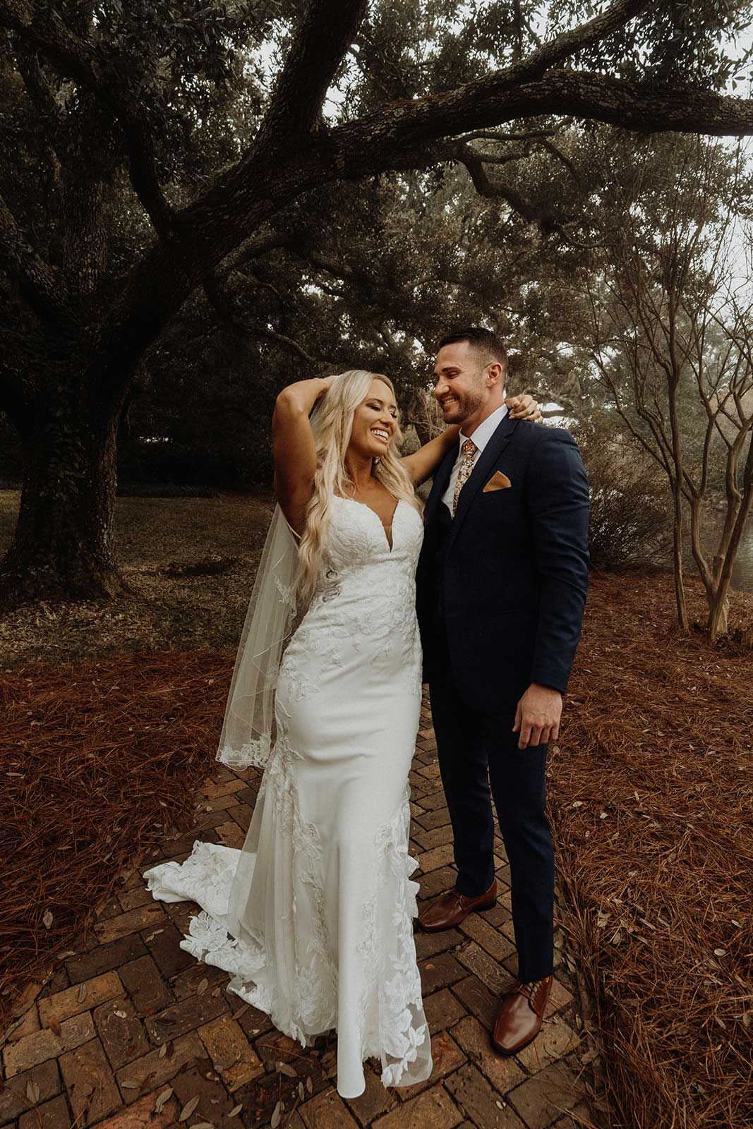 Bride and groom pose on a brick path, she in a white gown, he in a blue suit, under a tree.