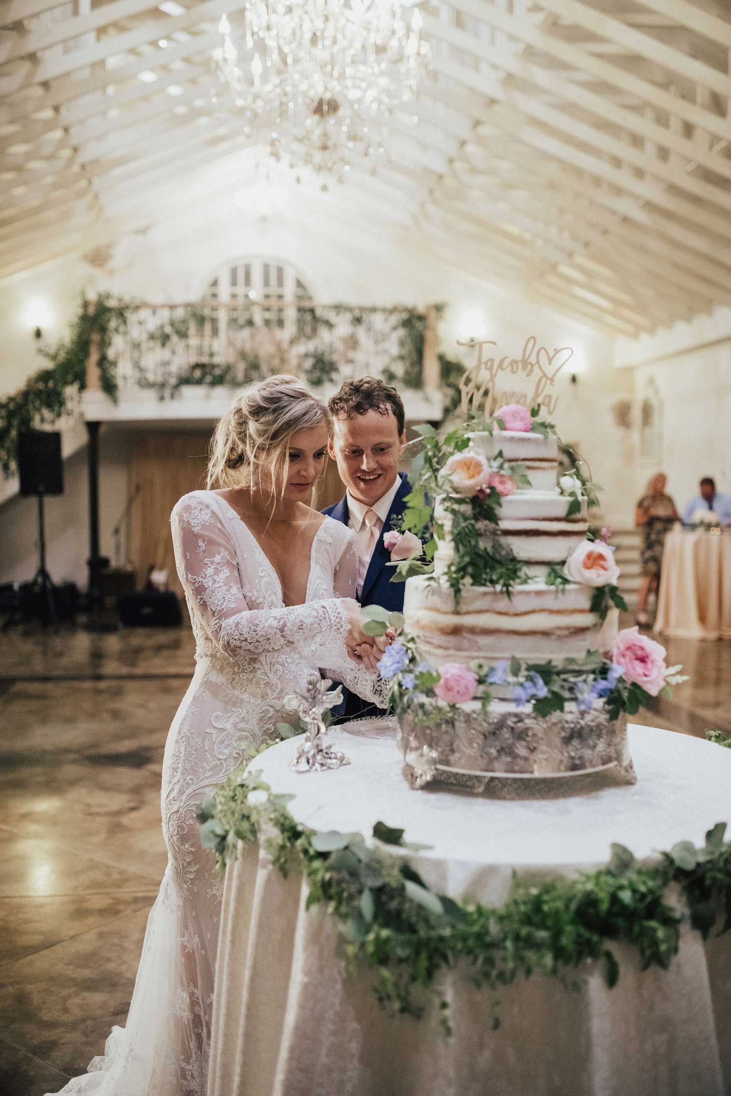 Couple cuts tiered wedding cake, decorated with flowers, in a warmly lit reception hall.