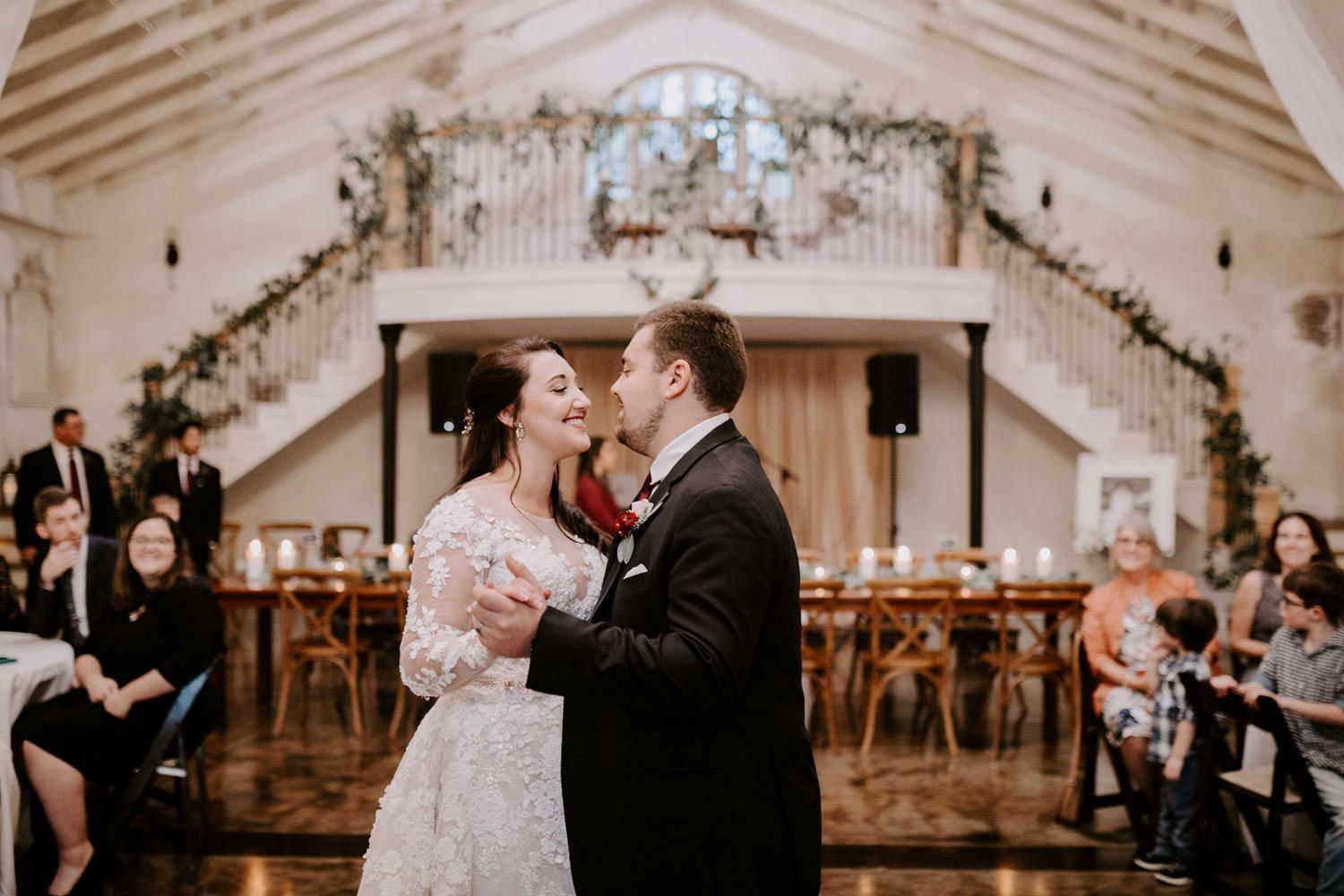 Newlyweds dance at reception, wooden tables and stairs with floral garland behind them.