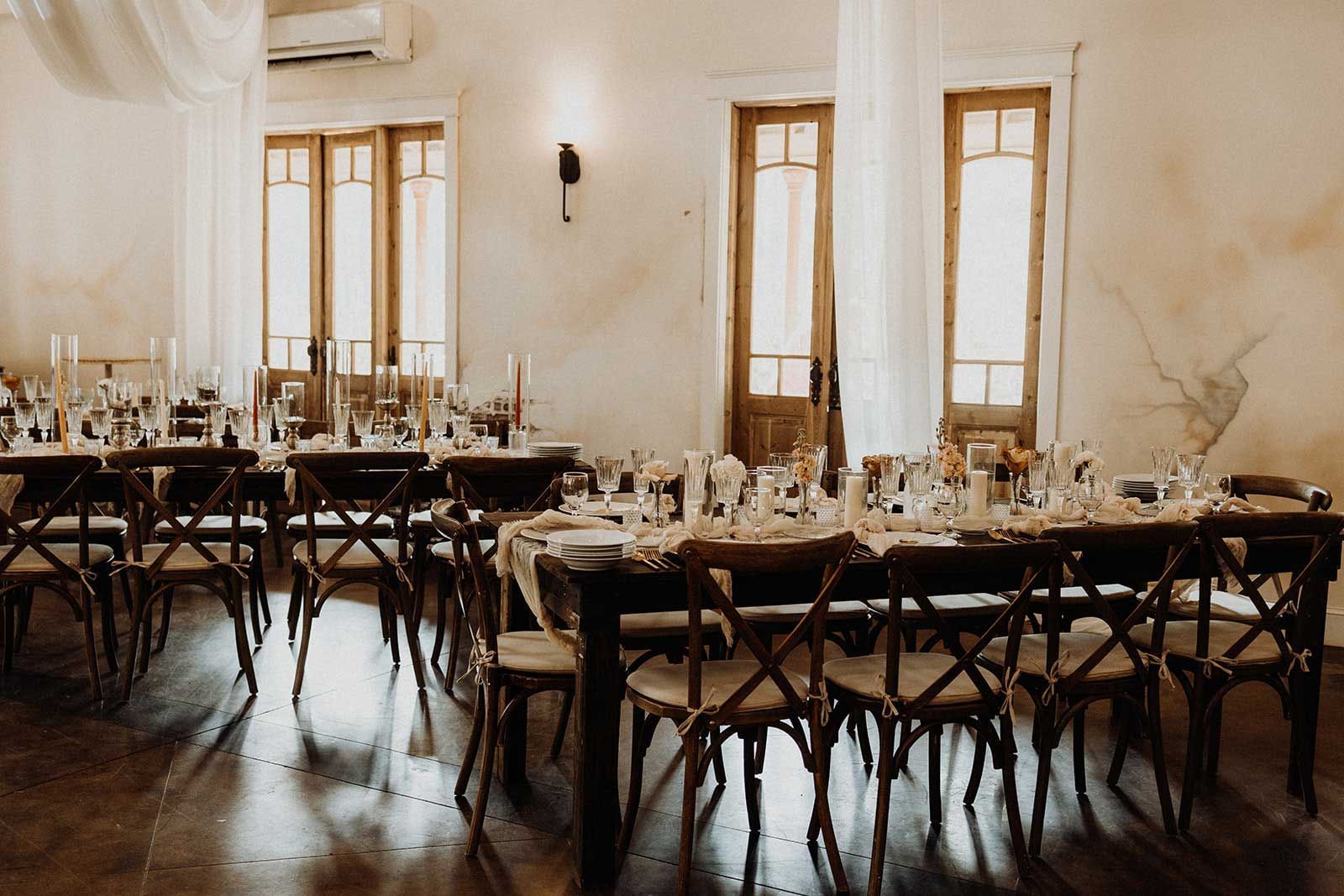 Wedding reception dining area with long wooden tables set for guests; rustic chairs, floral centerpieces.