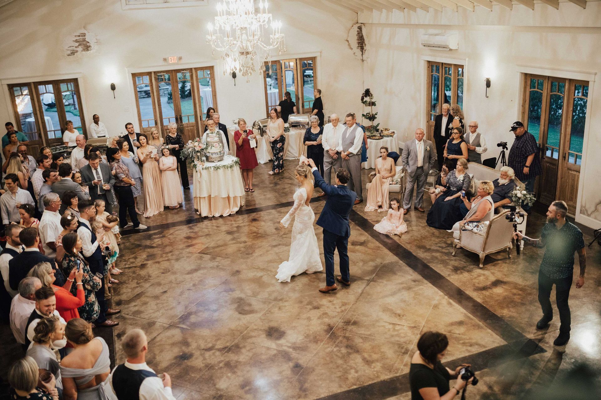 Newlyweds dance surrounded by guests in a reception hall with a chandelier.