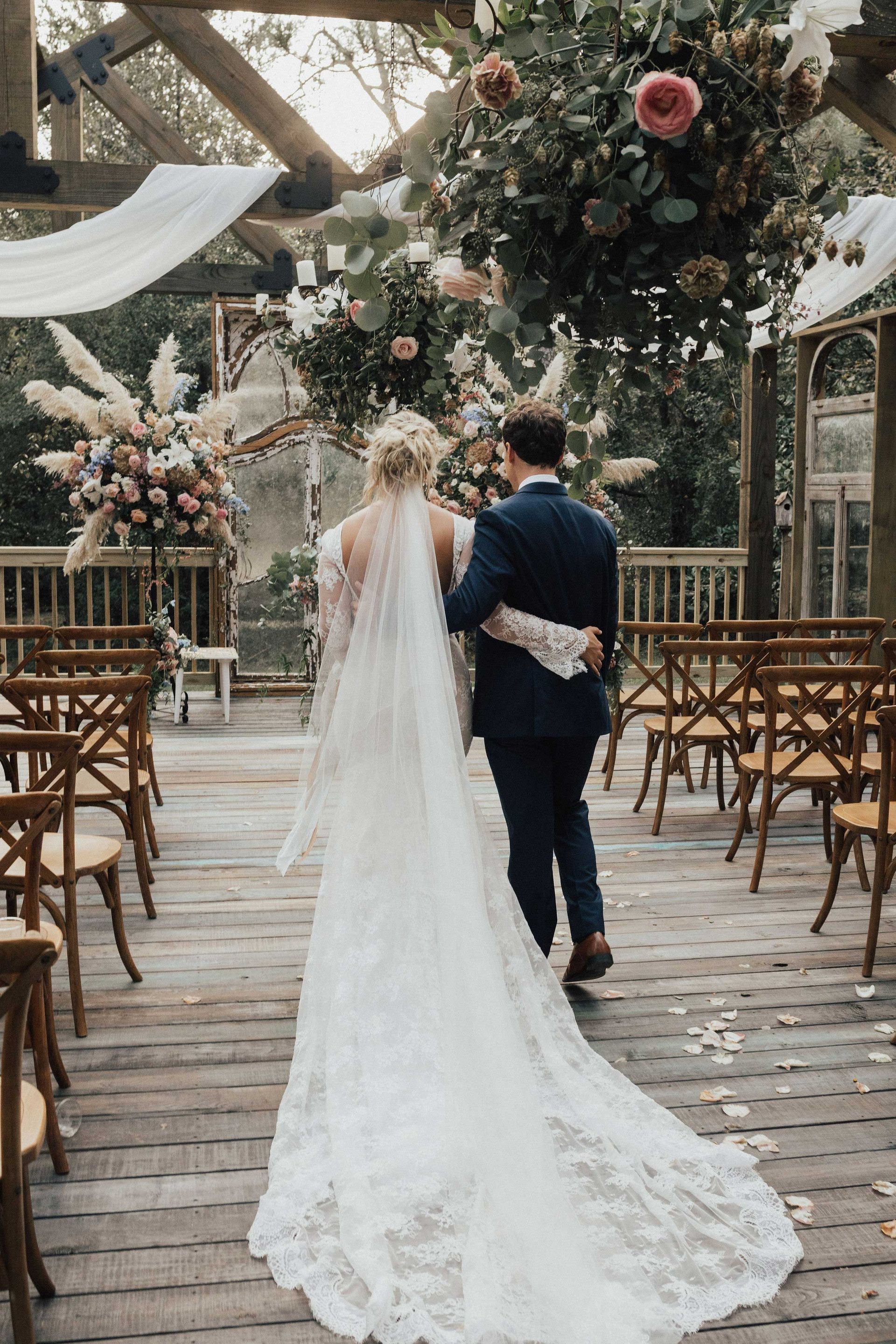 Bride and groom walking down an aisle, floral arch above, outdoor wedding venue.