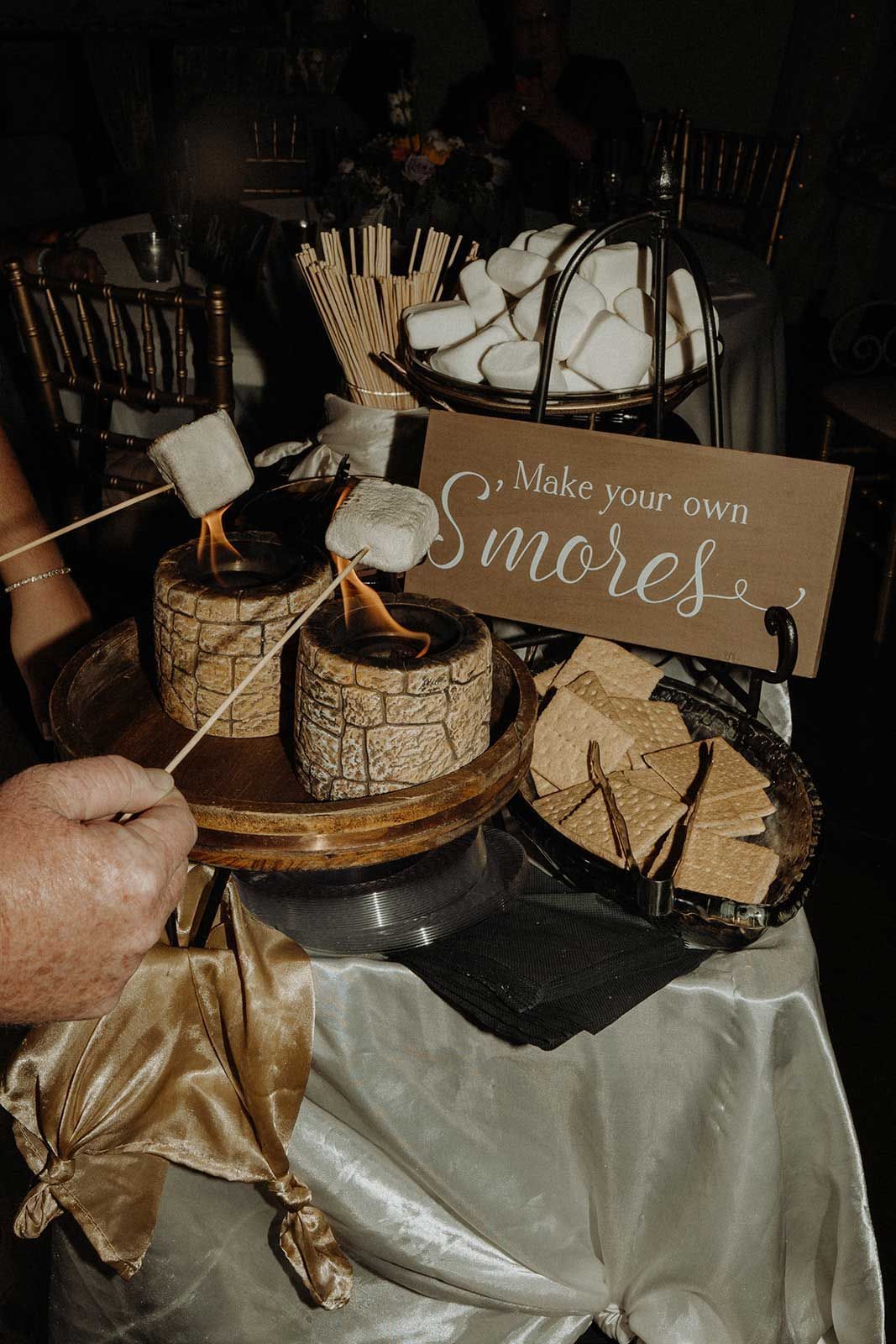 S'mores station: small fire pits, marshmallows roasting on sticks, graham crackers, sign.