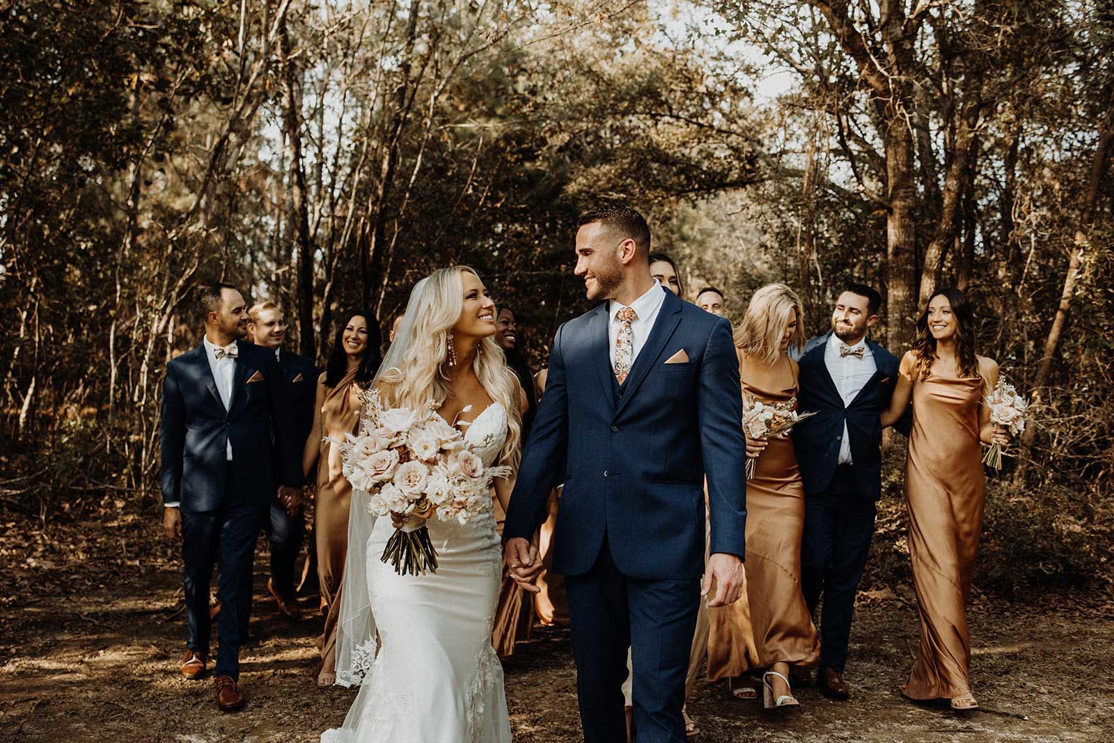 Wedding party walking in a forest. Bride and groom holding hands, smiling. Bridesmaids in gold dresses. Groomsmen in blue suits.