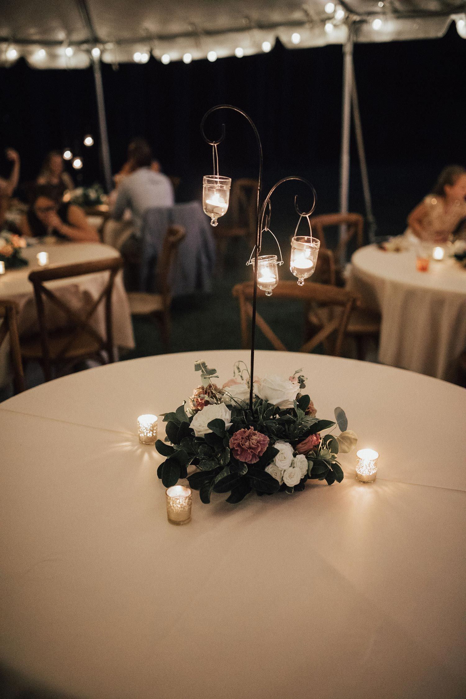 Wedding reception table with floral centerpiece, candles, and hanging lanterns under a tent.