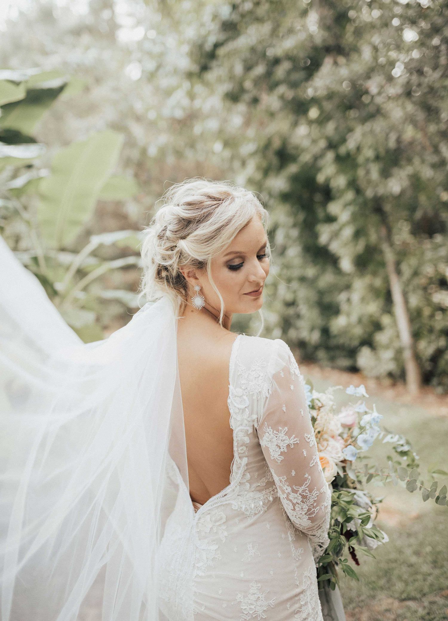 Bride in a white, open-back wedding dress, hair up, holding bouquet, veil flowing in an outdoor setting.