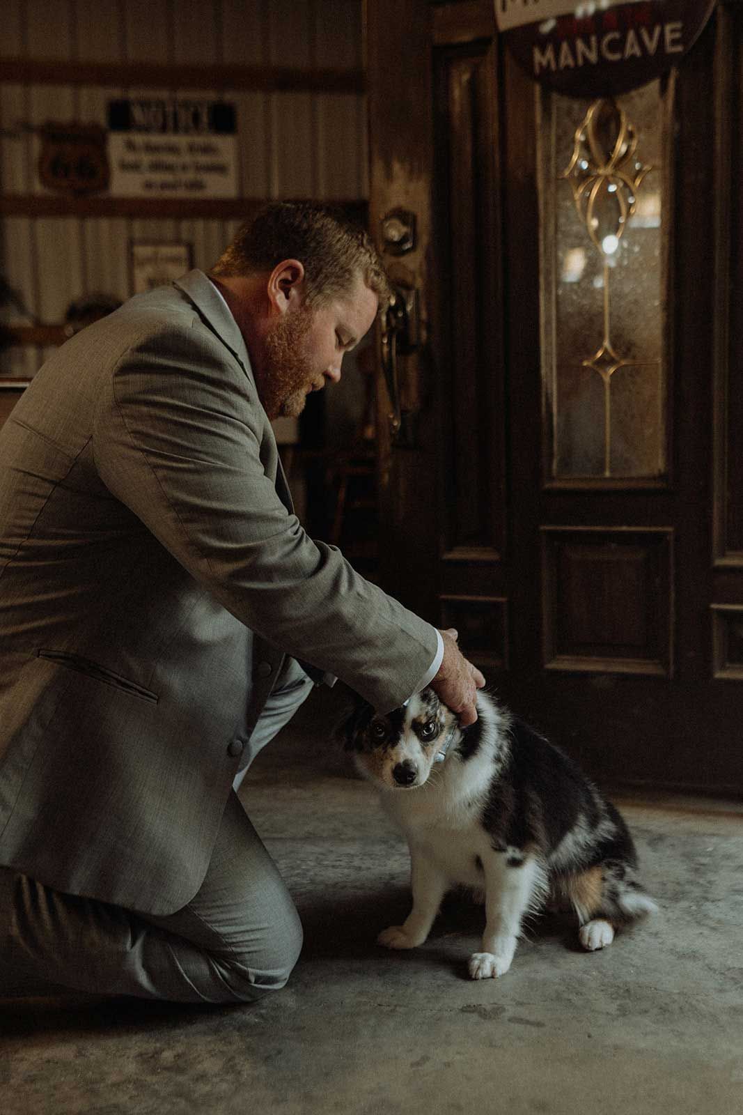 Man in gray suit kneels, petting a blue merle dog.