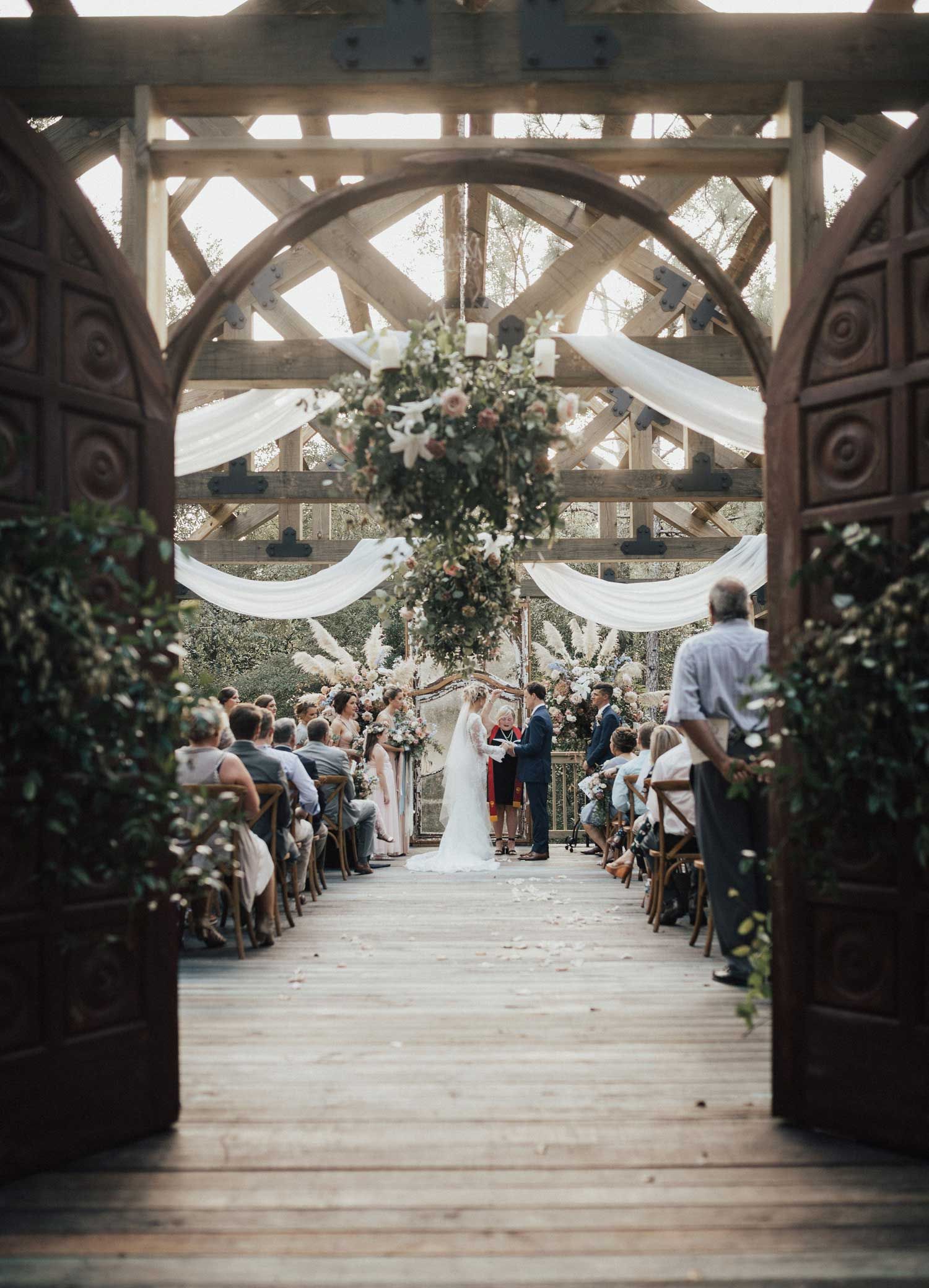 Wedding ceremony outdoors, under a wooden arch. Couple at the altar; guests seated on either side.