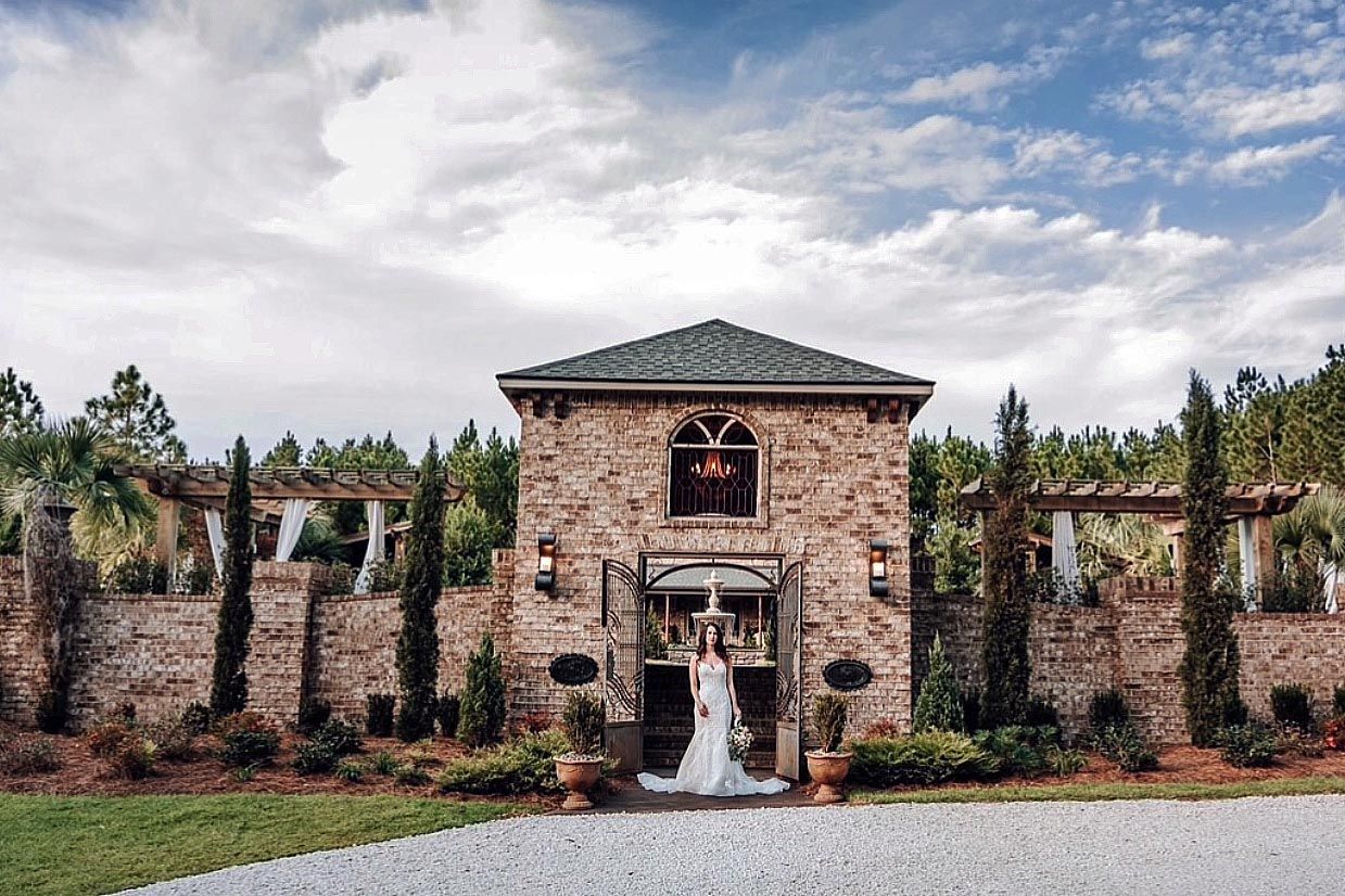 Bride in a wedding dress stands at the entrance of a brick building with a green roof, blue sky backdrop.