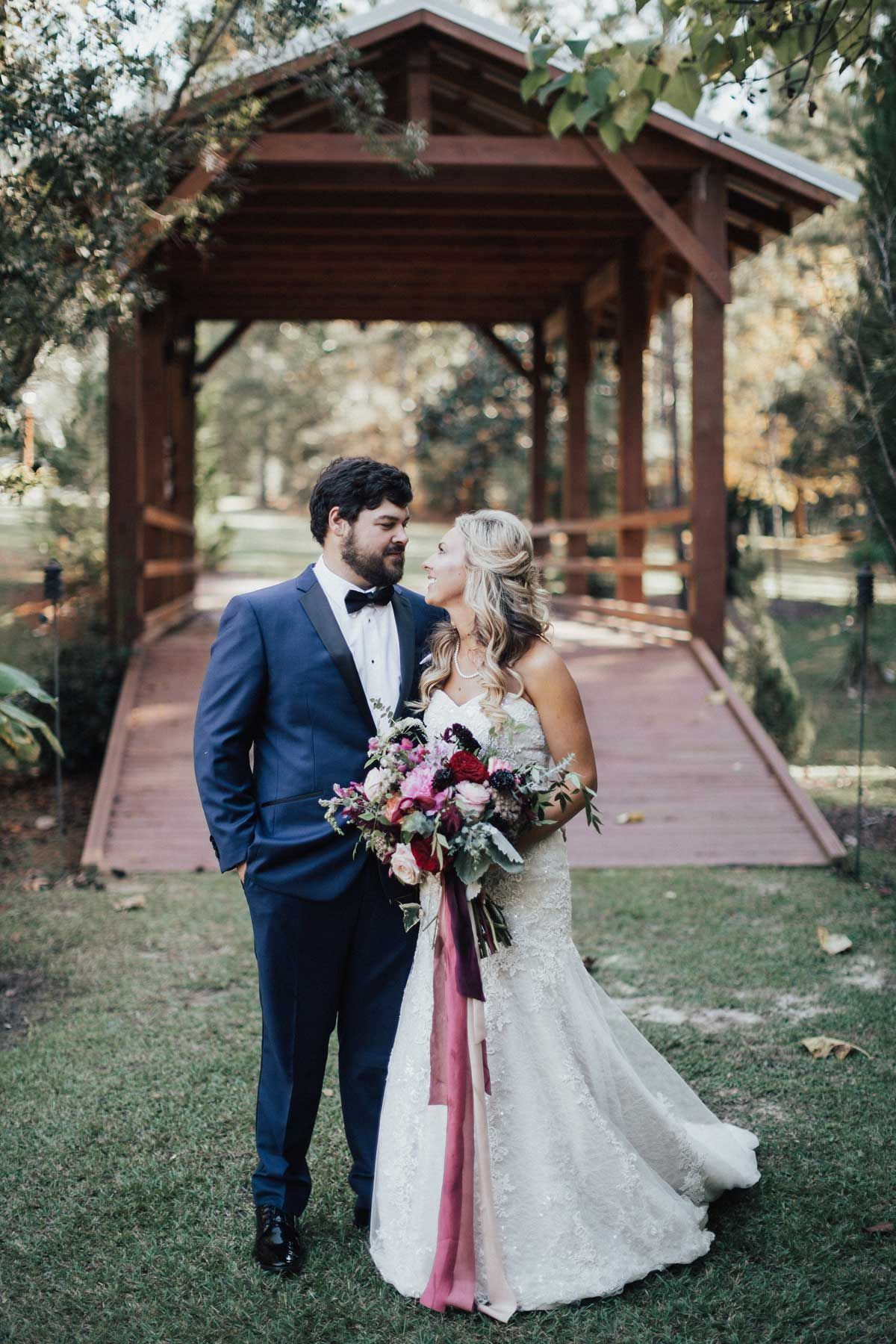 Wedding couple stands in front of a wooden bridge. The groom in a blue suit, the bride in a white dress, and they are holding a bouquet of flowers.
