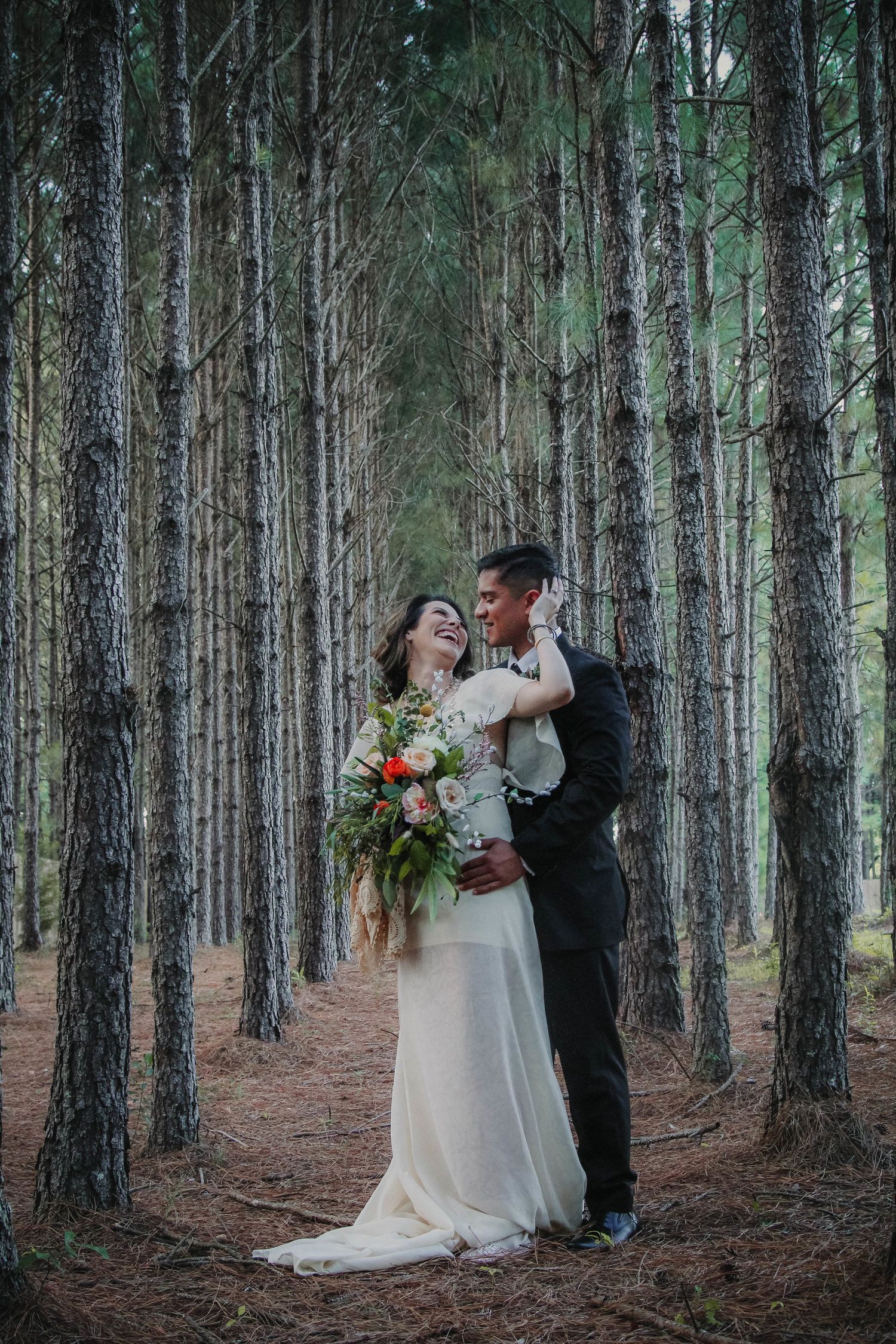 Couple embracing in a forest. Woman in white dress holds bouquet; man in a suit. Smiling, standing among trees.