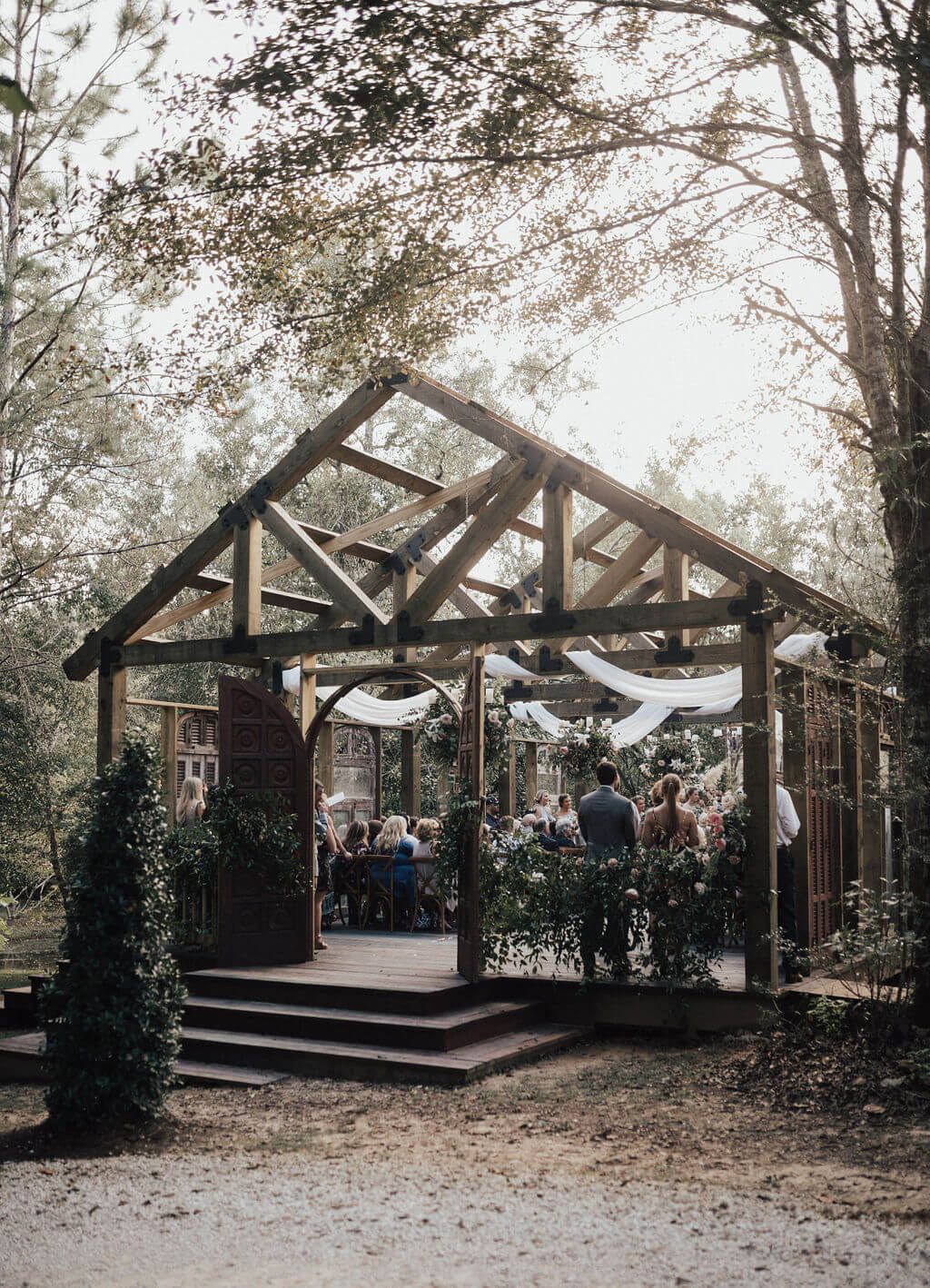 Wooden outdoor wedding structure in a forest. Guests seated under a canopy with draped fabric and greenery.