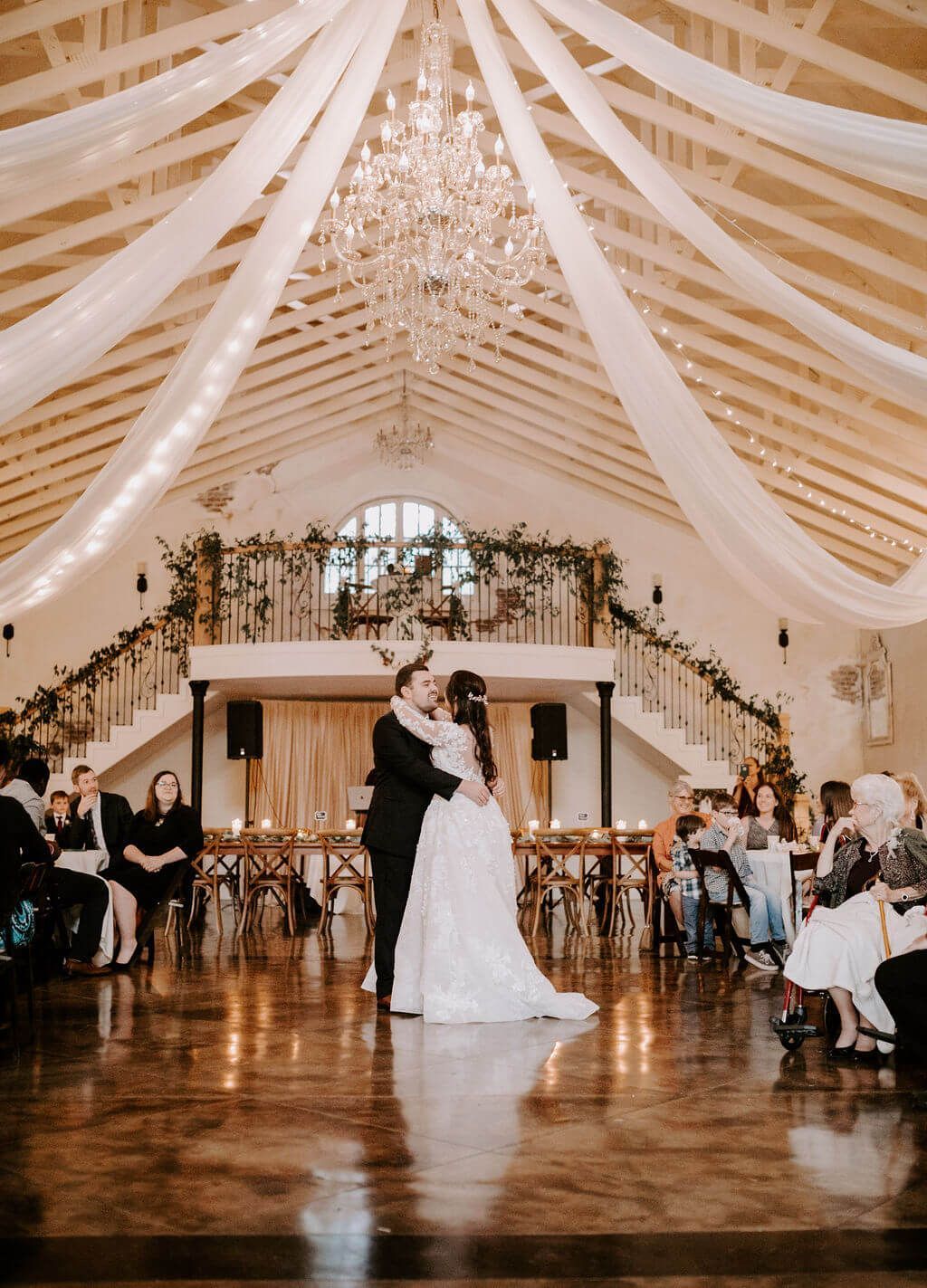 Couple dancing at a wedding reception, inside a decorated hall with draped fabric and a chandelier.
