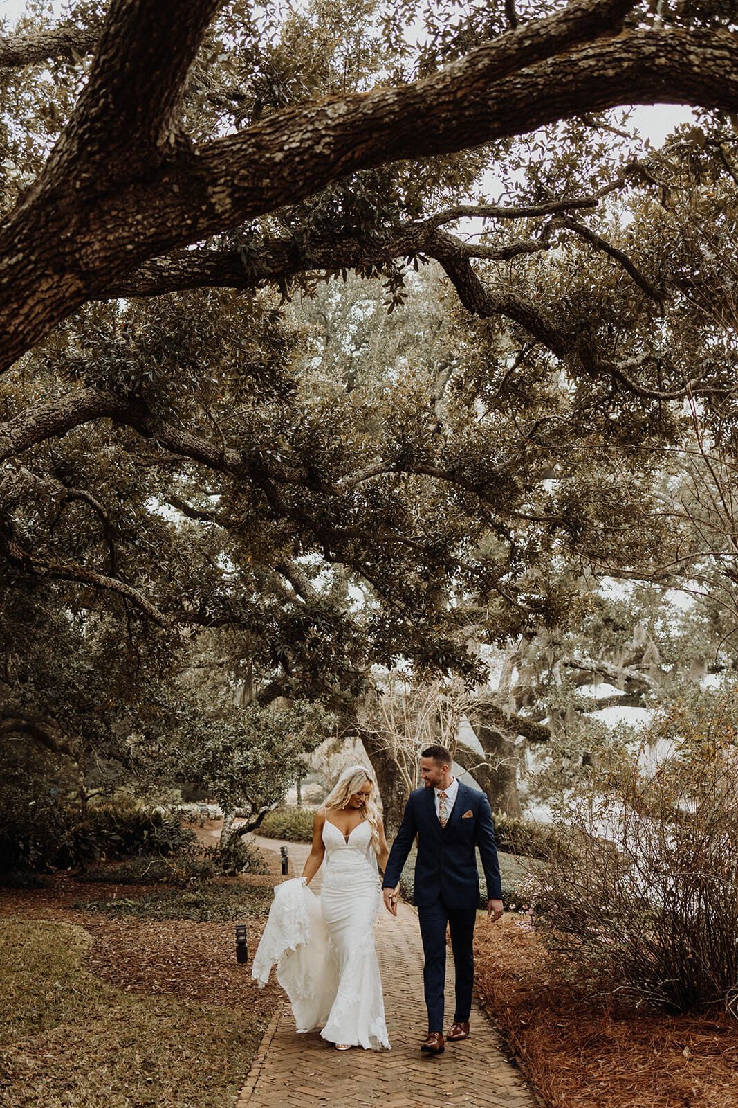 Bride and groom walk hand-in-hand on stone path under large tree with lake visible in the distance.