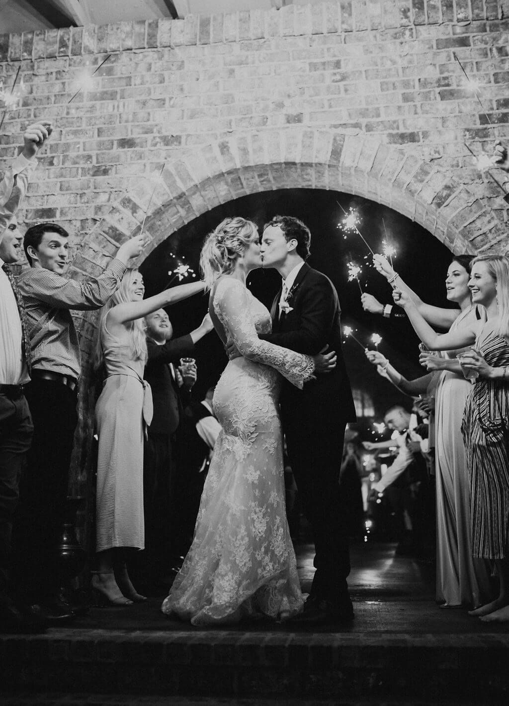 Couple kissing under brick archway as guests hold sparklers.