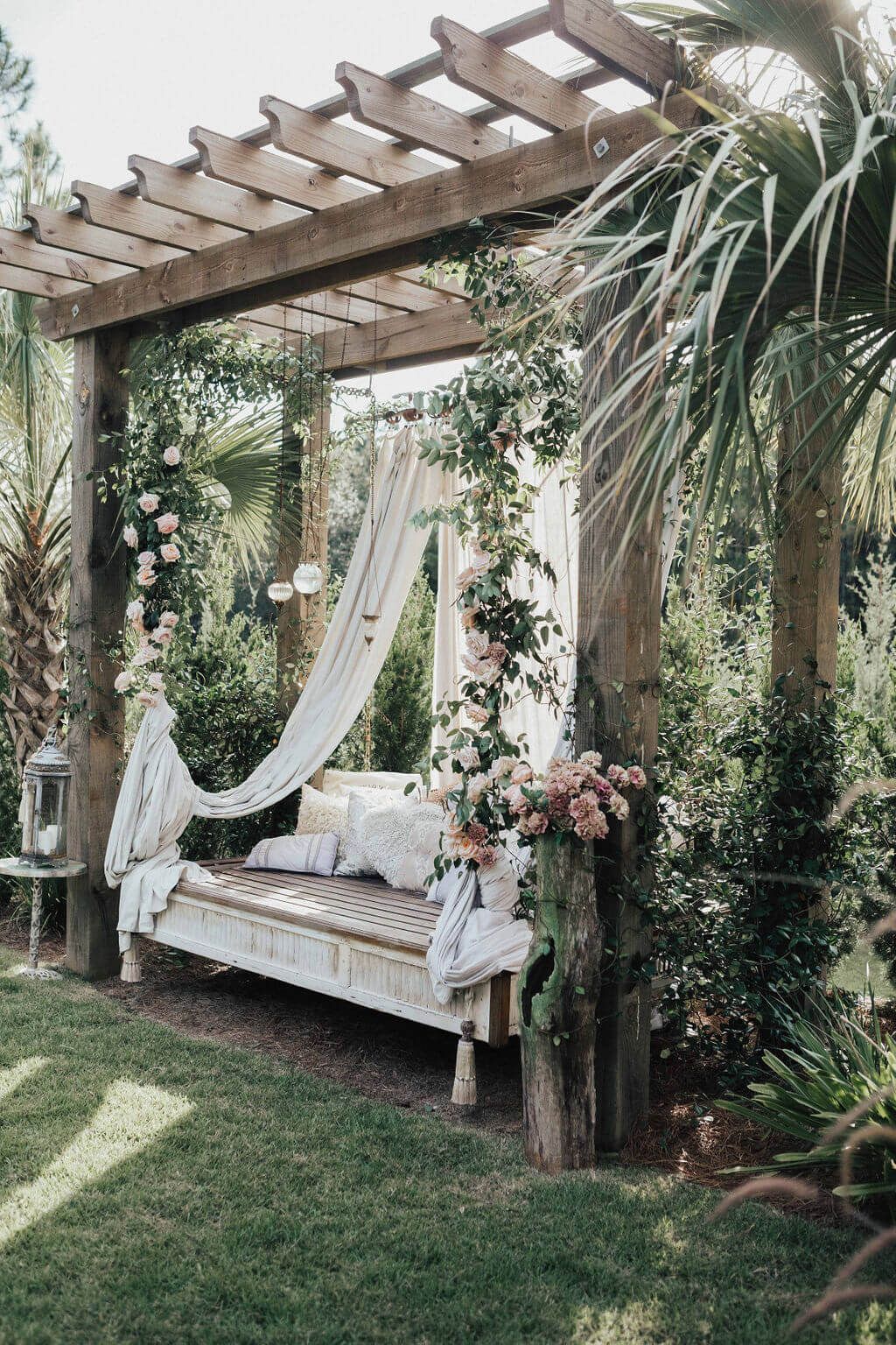 Outdoor wooden pergola with daybed, decorated with flowers and draped fabric, in a garden setting.