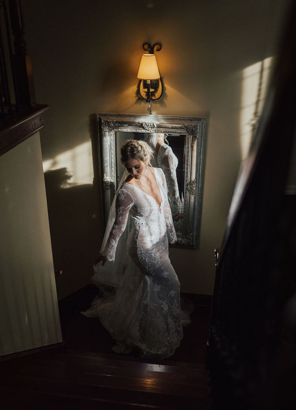 Bride in lace wedding dress on stairs, backlit by sunlight, near a mirror.