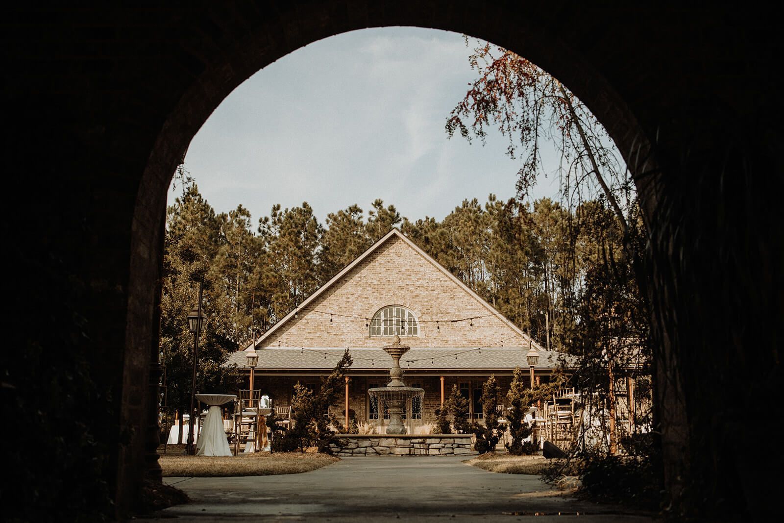 A light-colored building with a peaked roof is framed by an archway, trees, and sky.
