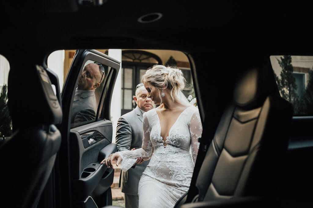 Bride exiting a black car, assisted by a man in a suit, at a building entrance.