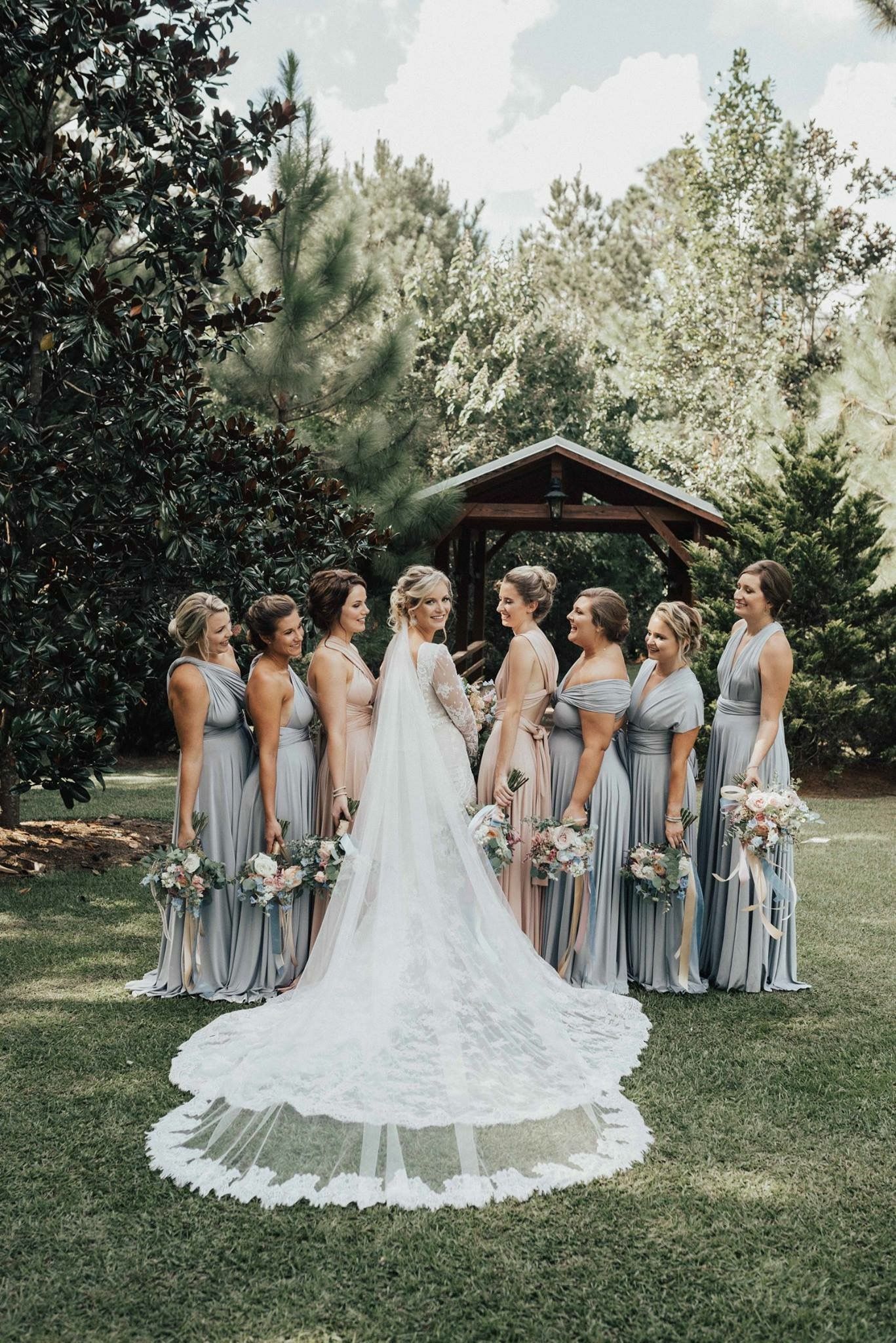 Bride in long veil with bridesmaids in blue dresses outdoors near an arbor.